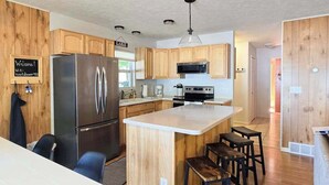 A modern kitchen with wooden cabinets, a stainless steel refrigerator, a microwave, and a central island with stools. - Glen Lake Home w/private beach frontage and dock (Empire)