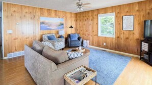 A living room with a sofa, a coffee table, a blue rug, wooden walls, and a ceiling fan. - Glen Lake Home w/private beach frontage and dock (Empire)
