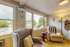 Une pièce dotée de grandes fenêtres, d’un fauteuil à carreaux, d’un fauteuil beige, d’une petite table recouverte d’une nappe rouge et blanche, et d’une lampe fixée au mur.