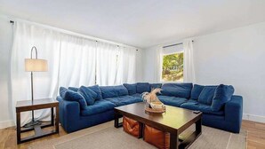 Un soggiorno con un divano angolare blu, un tavolino in legno, una lampada da terra e vista su del verde all'esterno.