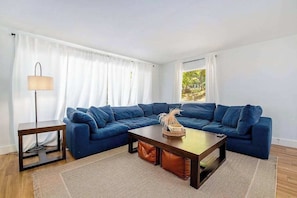 A living room with a blue sectional sofa, a wooden coffee table, a floor lamp, and a view of greenery outside. - Comfortable home in a great location with enclosed yard, family room, & kayaks (Frankfort)
