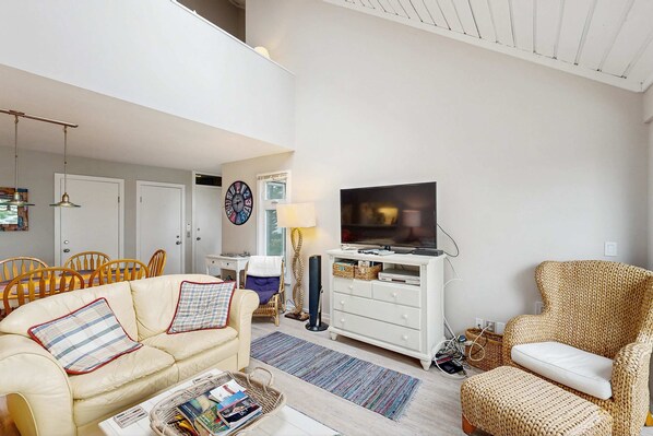 A spacious living room with a sofa, armchairs, a television, and a dining area with chairs and a table. - Beautiful beachfront townhouse with deck, water views, community firepit (Glen Arbor)