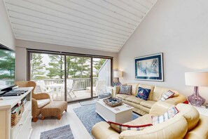 A spacious living room with a large sliding glass door leading to an outdoor deck, a comfortable sofa set, a coffee table, and a painting on the wall. - Beautiful beachfront townhouse with deck, water views, community firepit (Glen Arbor)