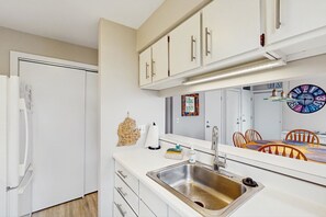 A kitchen with white cabinets, a stainless steel sink, a refrigerator, and a large mirror reflecting a dining area. - Beautiful beachfront townhouse with deck, water views, community firepit (Glen Arbor)