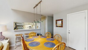 Un coin repas avec une table jaune, six chaises et une cuisine en arrière-plan.