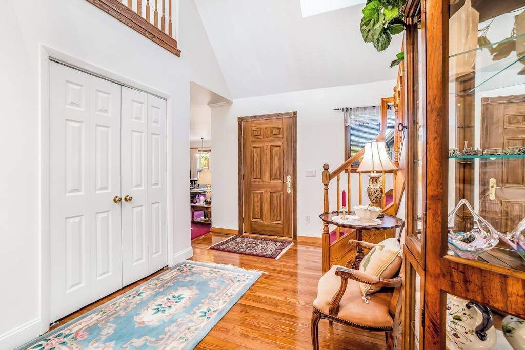 A hallway with a wooden floor, a white door, a wooden door, a chair, a table with a lamp, and a glass cabinet with decorative items.