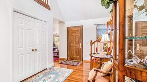 A hallway with a wooden floor, a white door, a wooden door, a chair, a table with a lamp, and a glass cabinet with decorative items.