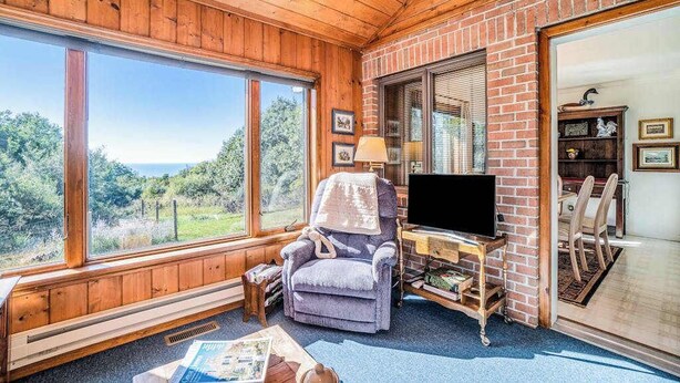 A cozy room with a large window, a television on a wooden stand, a chair, and a bookshelf.