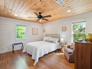 A bedroom with a bed, a ceiling fan, a nightstand, a lamp, a chair, and a window with a view of greenery.
