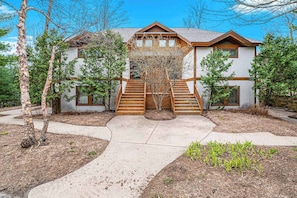 A two-story house with a wooden staircase leading to the front door, surrounded by trees and a landscaped garden.