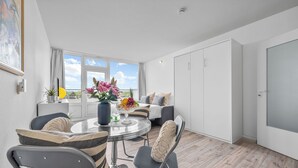 A modern living room with a round dining table, comfortable seating, a large window with a view, and a sliding door. - Cozy Holiday Apartment with Limited Sea View near the Beach in Wendtorf (Wendtorf)