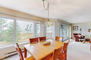 A dining area with a wooden table and chairs, a chandelier, and large windows with a view of trees. - Lovely Lake Michigan beachfront condo with deck, washer/dryer, fireplace, views (Glen Arbor)