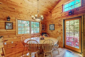 A wooden cabin interior with a dining table, chairs, a chandelier, and large windows.