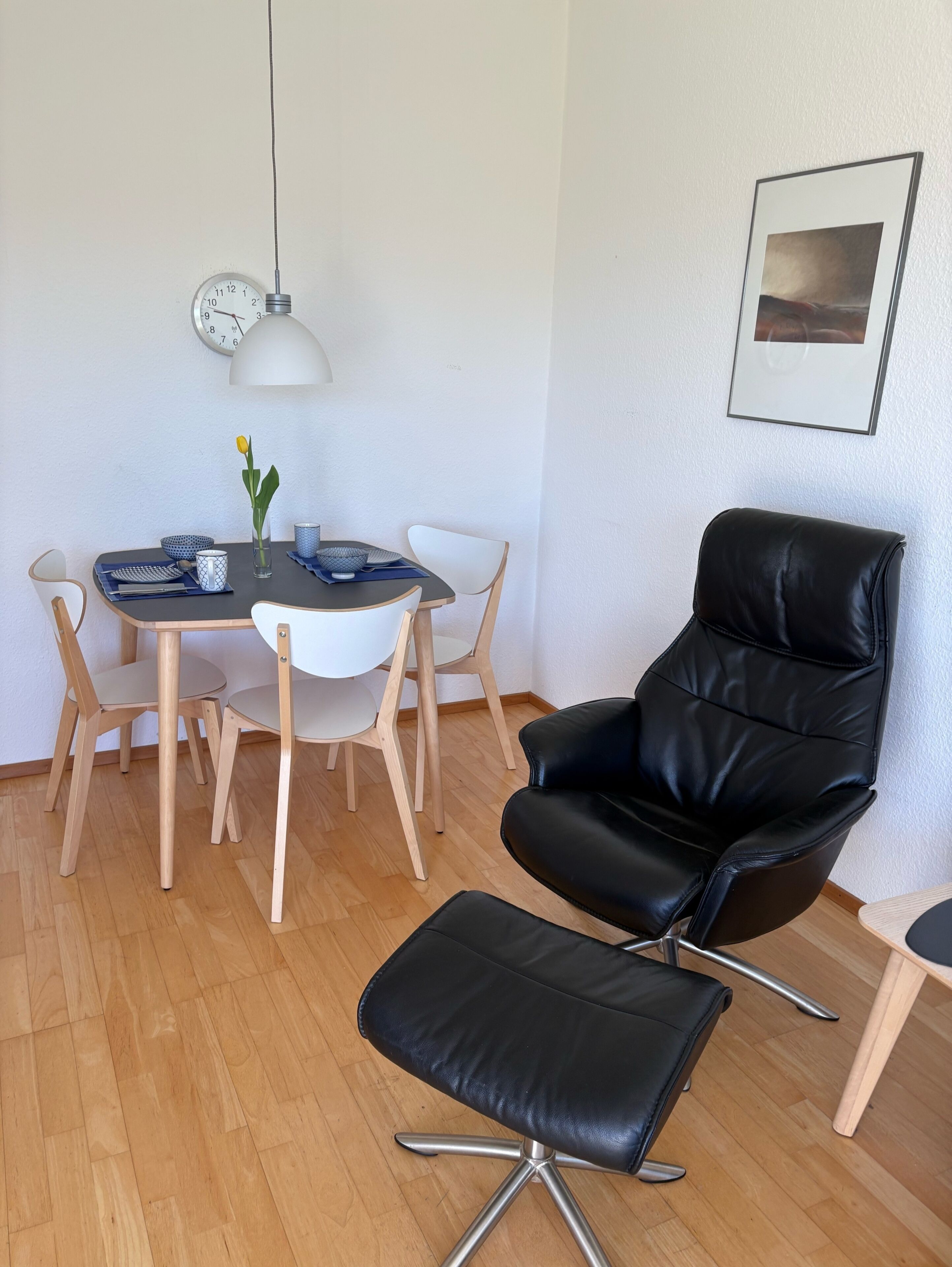 Et spiseområde med et rundt bord, stole, en sort læderstol, en lille skammel, et vægur og et indrammet billede.