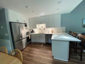 A modern kitchen with white cabinets, stainless steel appliances, a wooden dining table, and a large island. - River-view condo with fireplace, deck, washer/dryer, and community firepit (Glen Arbor)
