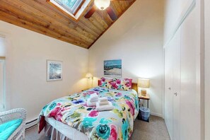 A bedroom with a wooden ceiling, a bed with floral bedding, a nightstand with a lamp, and a window with a view. - River-view condo with fireplace, deck, washer/dryer, and community firepit (Glen Arbor)