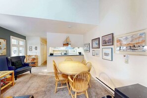 A living room with a dining table, chairs, a sofa, and a kitchen area with a hanging light fixture. - River-view condo with fireplace, deck, washer/dryer, and community firepit (Glen Arbor)
