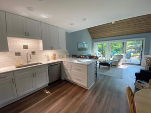 A modern kitchen with white cabinets, a wooden floor, and a dining area with a view of the outdoors. - River-view condo with fireplace, deck, washer/dryer, and community firepit (Glen Arbor)