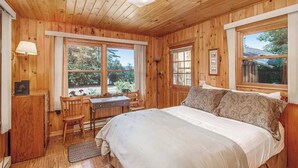 Ein Zimmer in einer Holzhütte mit einem Bett, Schreibtisch, Stuhl und großen Fenstern.
