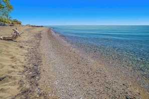 A pebbly beach with clear water, a driftwood piece, and a clear sky.