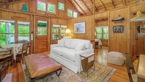 A wooden cabin interior with a white sofa, dining table, and chairs, wooden walls, and large windows. - Cabin-style cottage w/ full kitchen, wood-burning fireplace, and full WiFi (Frankfort)