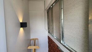 Um corredor estreito com um aplique na parede, uma cadeira de madeira e uma janela com moldura de metal.