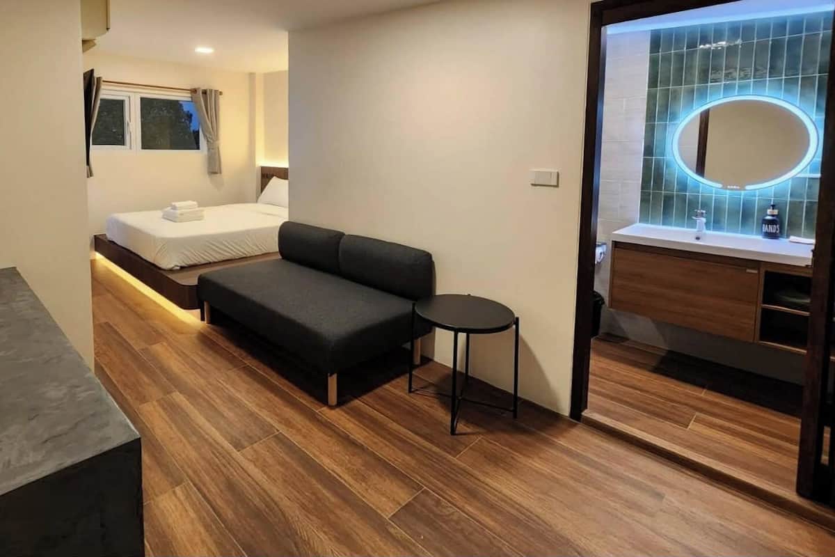A modern hotel room with a bed, a sofa, a small table, a mirror, and a bathroom with a vanity.