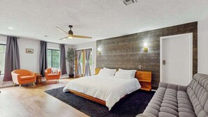 Ett modernt sovrum med en stor säng, två stolar, en soffa, en takfläkt och en vägg med träpanel som ger rummet en accentfärg.