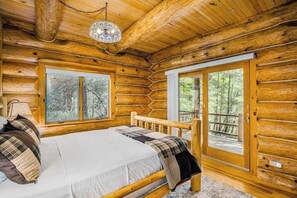 A log cabin bedroom with a bed, two windows, a door, and a chandelier.