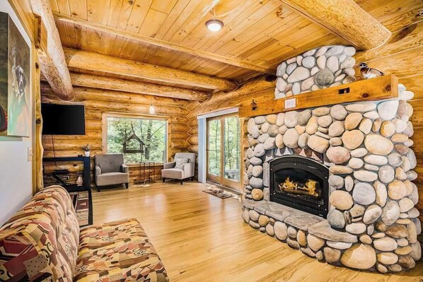 A log cabin interior with a stone fireplace, wooden ceiling beams, and a sofa.