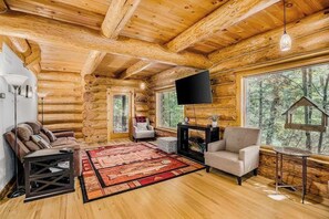 A log cabin interior with a TV, sofas, a fireplace, and large windows.