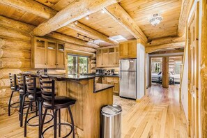 Ein rustikales Blockhaus-Interieur mit Küchenbereich, ausgestattet mit Holzsärchen, einem Edelstahl-Kühlschrank und einer Bar mit Hockern.