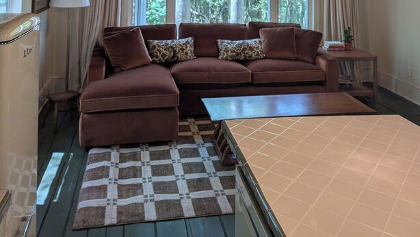 Ein gemütliches Zimmer mit Holzbalkendecke, großen Fenstern, einem braunen Sofa, einem gemusterten Teppich und einem Tisch.