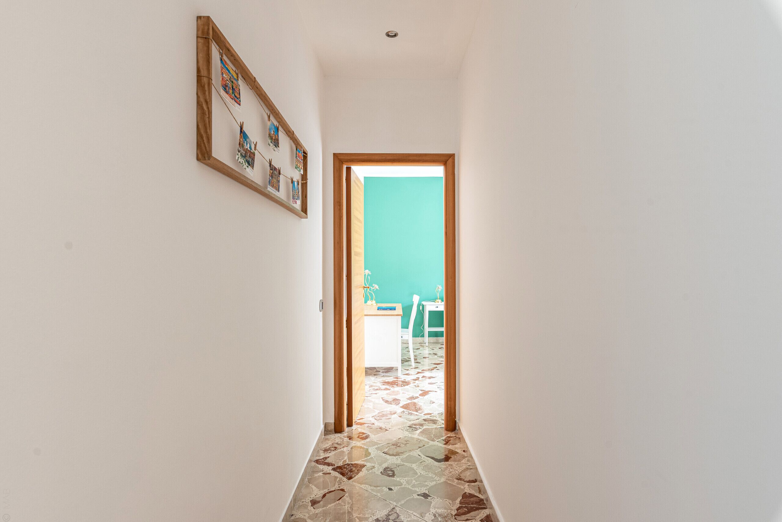 A narrow hallway with patterned flooring leading to a room with a teal wall and a white chair.