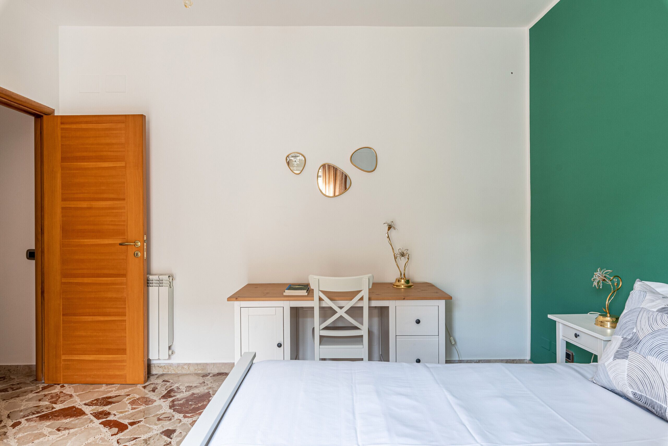 A bedroom with a bed, a desk with a chair, a radiator, and a door.