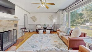 A living room with a fireplace, a ceiling fan, a large window, and a view of the outdoors. - Lovely home with hot tub, pool, two kitchens, enclosed backyard, firepit & grill (New Buffalo)