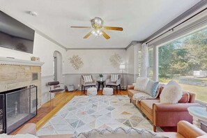 A living room with a fireplace, a ceiling fan, a large window, and a view of the outdoors. - Lovely home with hot tub, pool, two kitchens, enclosed backyard, firepit & grill (New Buffalo)