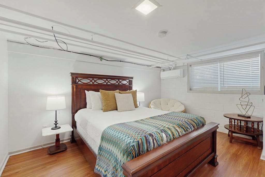 Een slaapkamer met een houten bed, witte wanden, een raam met zonwering, een nachtkastje met een lamp en een kleine ronde tafel met een decoratief object.