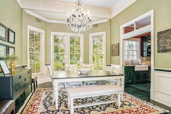 A dining area with a chandelier, a table with chairs, a bench, and a view of the outdoors through large windows. - Luxurious home with woodstove, fireplace, & screened porch - walk everywhere (Saugatuck)