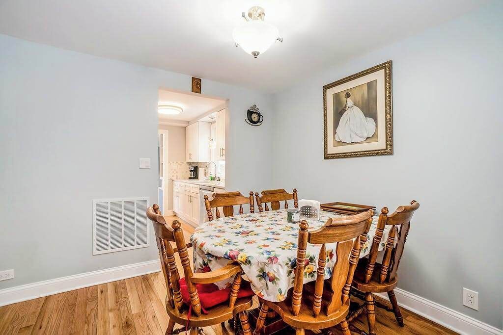 A dining area with a round table, wooden chairs, a floral tablecloth, a wall clock, and a framed picture.
