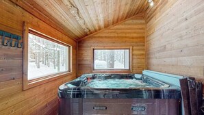 A wooden cabin interior with a hot tub, large windows, and a sloped ceiling. - Resort home with private indoor hot tub, game room, firepit, bikes, Ping-Pong (Thompsonville)