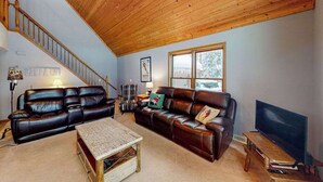 A living room with a wooden ceiling, a staircase, leather sofas, a coffee table, a TV, and a window with a view. - Resort home with private indoor hot tub, game room, firepit, bikes, Ping-Pong (Thompsonville)