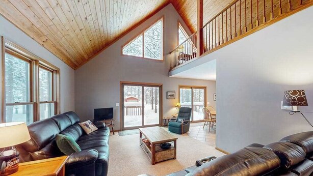 A spacious living area with a high ceiling, large windows, a leather sofa, a coffee table, and a balcony. - Resort home with private indoor hot tub, game room, firepit, bikes, Ping-Pong (Thompsonville)