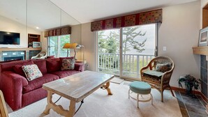 Ein Wohnzimmer mit einem roten Sofa, einem Holztisch, Korbstühlen, einem Fernseher und einem großen Fenster mit Blick auf Bäume.