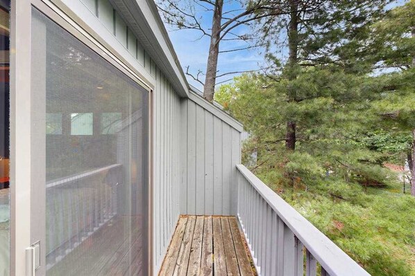 Una terraza de madera con un ventanal de vidrio que da a una habitación, rodeada de árboles.