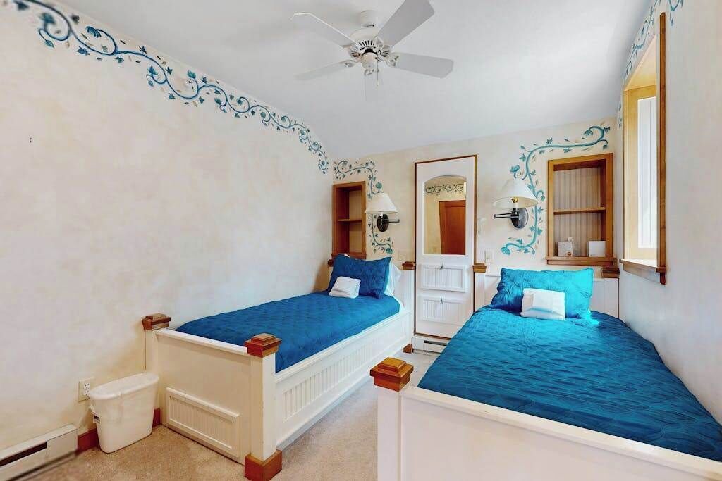 A bedroom with two beds, a ceiling fan, a door with a mirror, and a small trash bin.