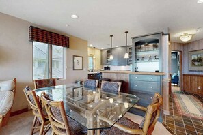 A dining area with a glass table and wicker chairs, a kitchen with blue cabinets and pendant lights, and a view of the outside through a window. - Lakefront condo with deck, gorgeous view, fireplace, & W/D (Glen Arbor)