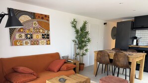 Sala de estar moderna com sofá, mesa de jantar e cadeiras. Há uma planta em vaso e uma obra de arte na parede.