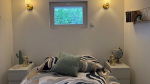 Quarto com uma cama, duas mesas de cabeceira, janela com vista para o verde, luminárias na parede e uma estante de livros.