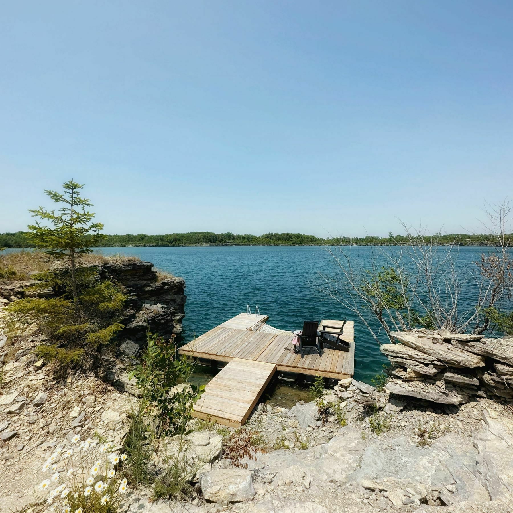 Un muelle de madera con sillas y una mesa se adentra en un lago, rodeado de terreno rocoso y vegetación escasa.